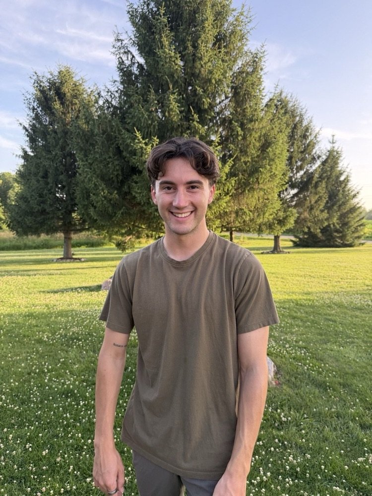 Drake Surach standing outside on a lawn in front of spruce trees at golden hour